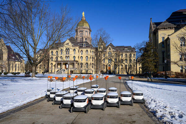 15 white and black autonomous delivery robots in a "V formation" in front of the Gold Dome of Notre Dame with snow on the ground