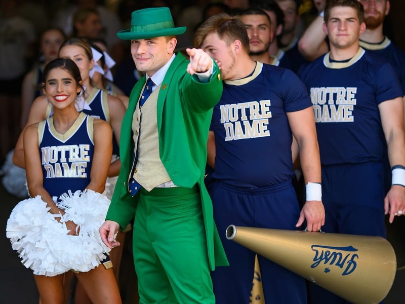 The ND Leprechaun dressed in green pointing at the camera. A group of male and female cheerleaders dressed in blue standing behind him.