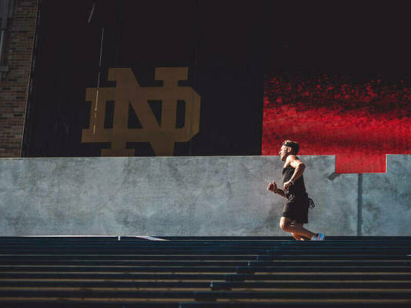 Man running in a row of the stadium stairs with the interlocking gold ND on a black background behind him