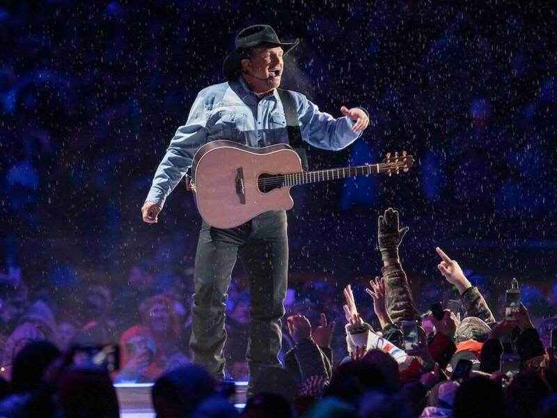 Garth Brooks standing on stage singing to a crowd of people with a guitar over his shoulder