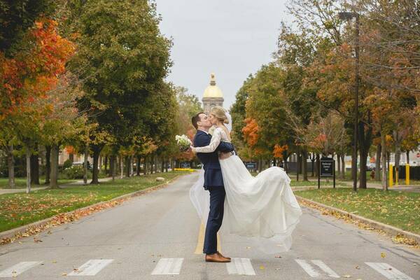 Groom lifting bride for a kiss on crosswalk with Golden Dome and fall foliage in background