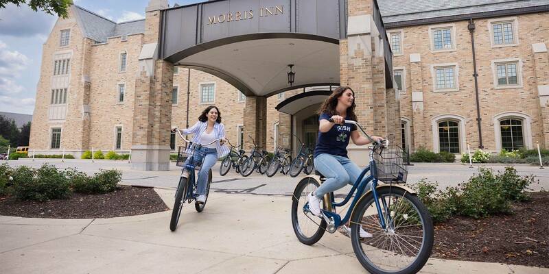 Two women riding bikes in front of the Morris Inn.