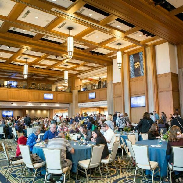 An event being held at Dahnke Ballroom inside of the Duncan Student Center. Circular tables with people around them fill the room. A stage sits at the front.