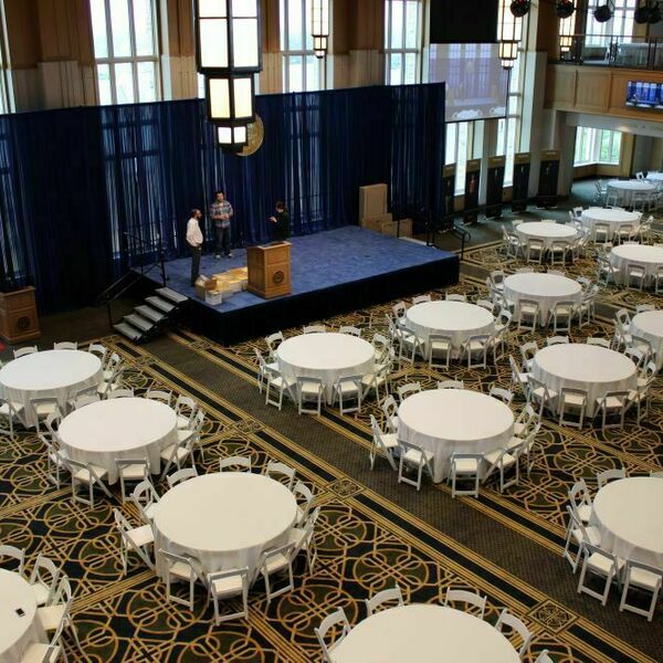 An event being set up in Dahnke Ballroom inside of the Duncan Student Center. Circular tables with white tablecloths and white chairs fill the room. A stage sits at the front of the room.