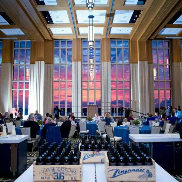 An event being held at Dahnke Ballroom inside of the Duncan Student Center. A pretty sunset with pink and purple colors is seen in the windows. Tables with blue tablecloths fill the space.