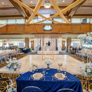 Downes Club event space decorated for a wedding. A DJ booth with white drapes in the back stands in front of a large white tile dancefloor. Long white tables with blue and white flowers line the sides of the room. A bride and groom table with a navy tablecloth sits at the front of the room.