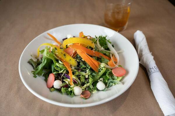 Salad in a white plate with arugula, yellow and orange peppers, mozzarella balls, next to a white rolled silverware on a brown paper tablecloth in Legends restaurant.