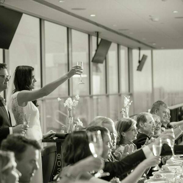 A bride and groom toast their glasses to many guests sitting at a table.