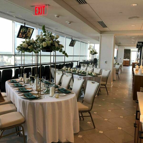 Circular tables with white tablecloths, green and gold place settings, and elaborate white and green flowers. The football stadium shines through the windows.
