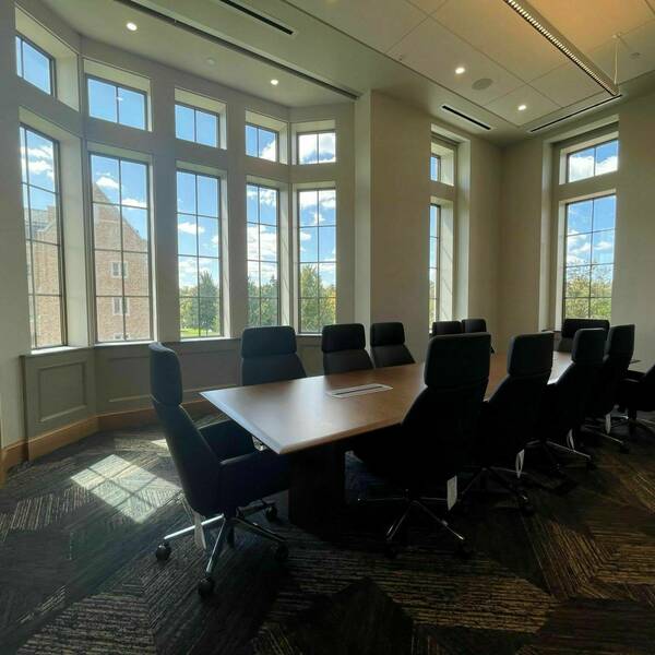Twelve black office chairs sit around a long rectangular, wooden table. Sunlight is beaming through multiple large windows.