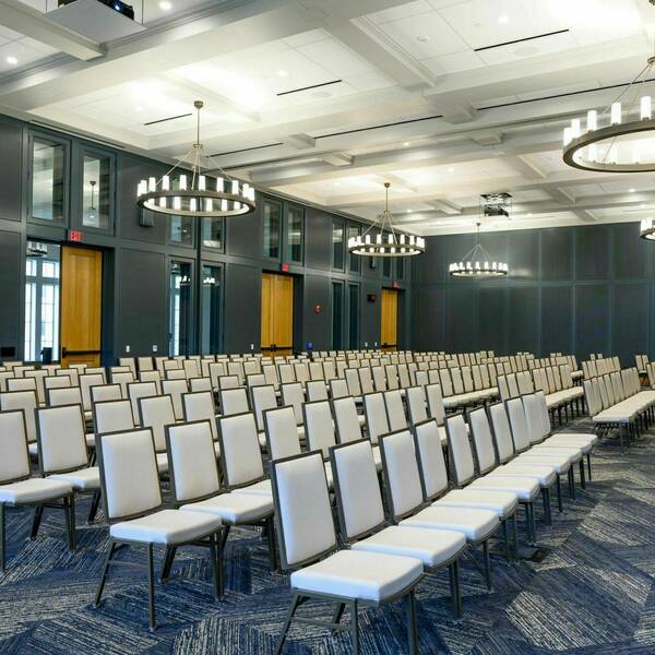 White leather chairs in long rows fill the room. A clear podium sits at the front. Large circular, modern chandeliers hang from the ceiling.