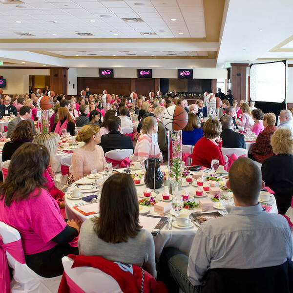 White tables with pink accents fill the space. People sit around decorated tables and listen to a speaker speaking on a small stage at the front of the room.