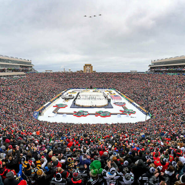 Notre Dame stadium filled with guests for the NHL Winter Classic. The field is covered in ice and shamrock structures. Planes fly above and the library can be seen in the distance.