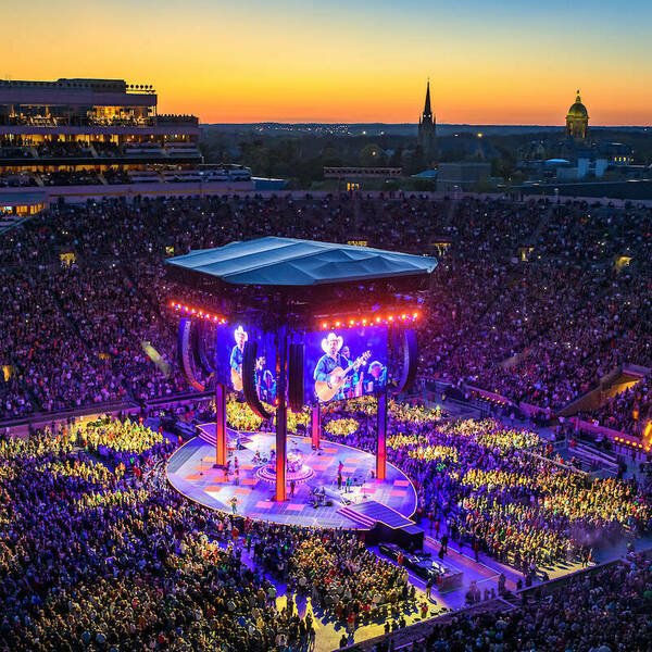 Notre Dame stadium filled with guests for the Garth Brooks concert. A big stage and screens sit in the field with guests surrounding it. Bright colors shine across the stadium.