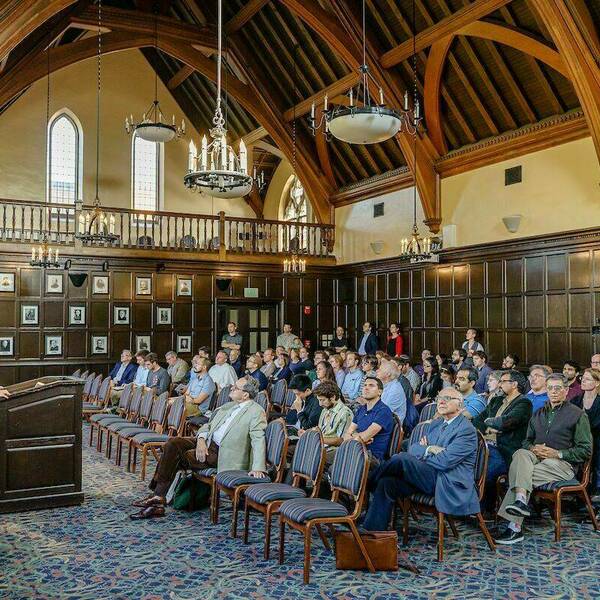 Rows of people sit in chairs listening to a speaker standing behind a podium at the front of the room. Wooden paneling fills the room and a second floor with large windows can be seen.