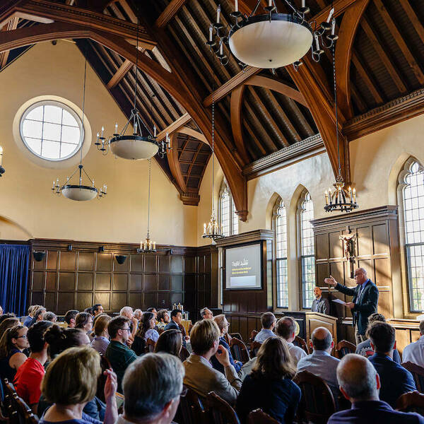 Rows of people sit and listen to a speaker at the front of the room. Wooden paneling fills the walls and elaborate curved wooden beams line the ceiling.