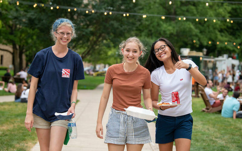 Three women students smiling and walking. Students gather for food trucks on the library quad.