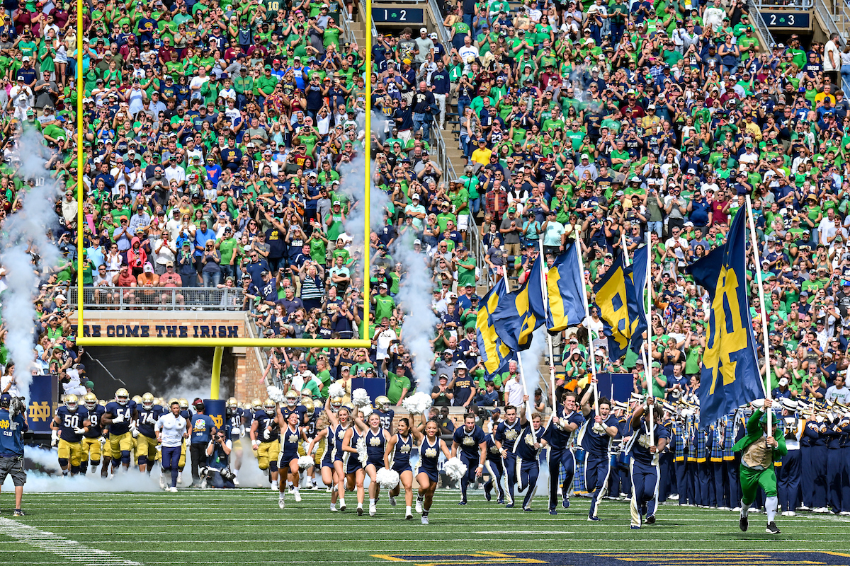 The football team runs out onto the football field. The cheerleaders holding large flags that spell Irish run in front of them. A stadium packed full of people can be seen in the back.