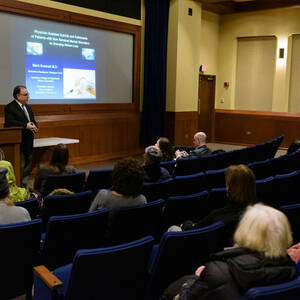 Students sitting in auditorium style seating listening to an adult speak at the front. A podium and projector screen fill the front of the room.