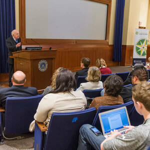 Students sitting in auditorium style seating listening to an adult speak at the front. A podium, projector screen, and signs fill the front of the room.