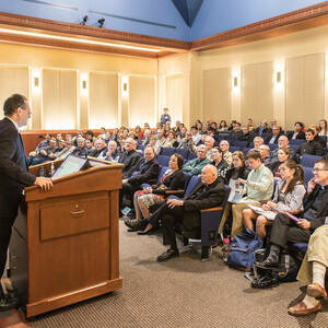 Rows of people sit in blue theater chairs to listen to a presenter at the front podium. Light colored walls and a blue ceiling decorate the room.