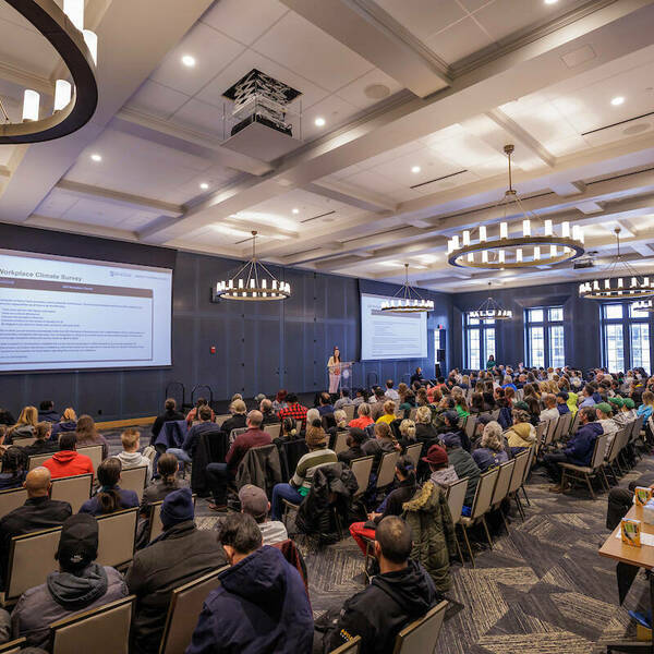 Large circular chandeliers hang from the ceiling and dark gray wood lines the walls. A stage sits at the front with a podium and a presenter. Large projector screens hang behind the presenter and long windows fill the side walls. Rows of people in chairs sit to watch.