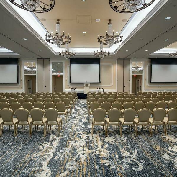 Lines of chairs fill the room. Navy and gold decorative carpet covers the floor. Crystal chandeliers hang from the ceiling, 3 large projectors hang at the front, and a small stage and podium sit at the front. Light tan walls and white framing fill the walls.