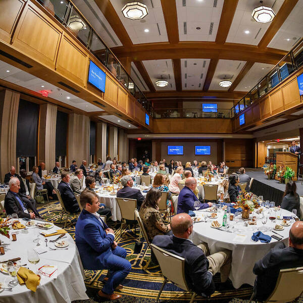 Event being held in Downes Club. Guests sit around white tables and listen to a speaker speaking on a stage at the front of the room. Wood paneling lines the ceiling, floor to ceiling windows line the back wall, and television screens hang along the walls.
