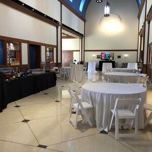 Circular white tables fill the atrium. Tan marble floors and wooden framing decorate the area. The building has a large dome like curvature for a ceiling and sections of it are painted vibrant blue. A long black table with a variety of drink options lines the edge of the room.