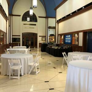 Circular white tables fill the atrium. Tan marble floors and wooden framing decorate the area. The building has a large dome like curvature for a ceiling and sections of it are painted vibrant blue. A long black table with a variety of drink options lines the edge of the room and a light up letters spell 