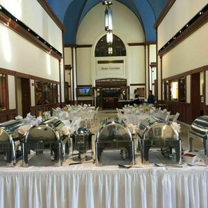 Circular white tables fill the area. an marble floors and wooden framing decorate the area. The building has a large dome like curvature for a ceiling and sections of it are painted vibrant blue. A long white table with multiple food options on it sits at the back of the room.