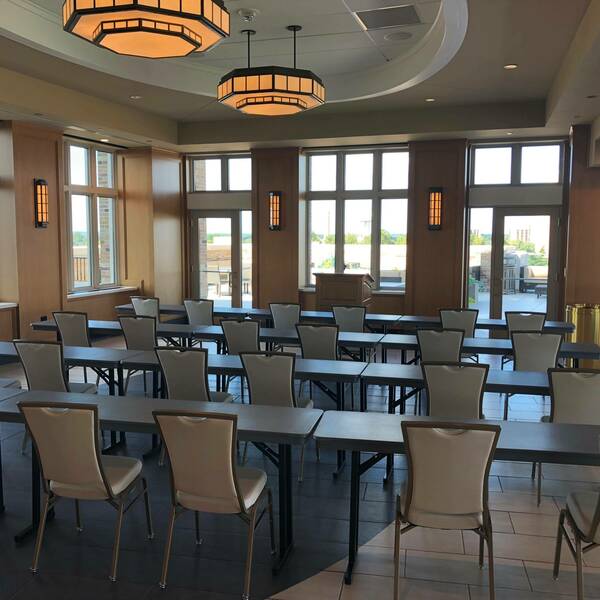 Rows of chairs and tables fill the space. Windows line the side of the room and a podium sits at the front.