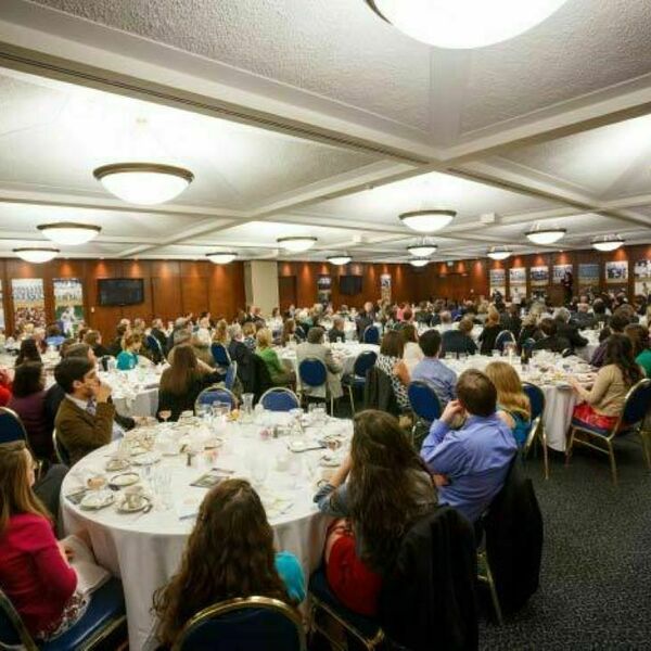 Groups of people sit at white circular tables for dinner. People are listening to a speaker talking at the front. Banners with different images line the walls of the room. Wood paneling fills the walls of the space.