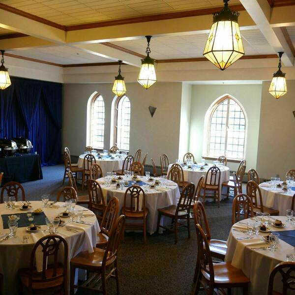 Large windows line the walls and vintage lights hang from the ceiling. Circular tables with traditional wooden chairs fill the room and a long table with food options sits at the front.