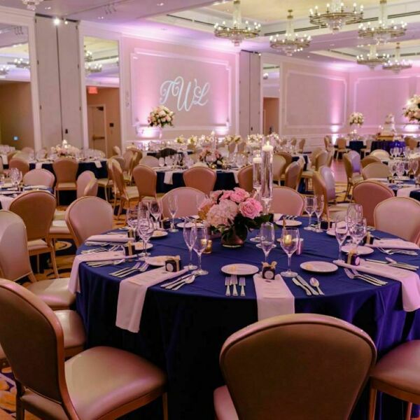 Circular tables with navy blue tablecloths and flowers fill the room. Settle pink lighting and crystal chandeliers give the room an elevated feel. The bride and groom's initials are projected on the wall above their table at the front of the room.