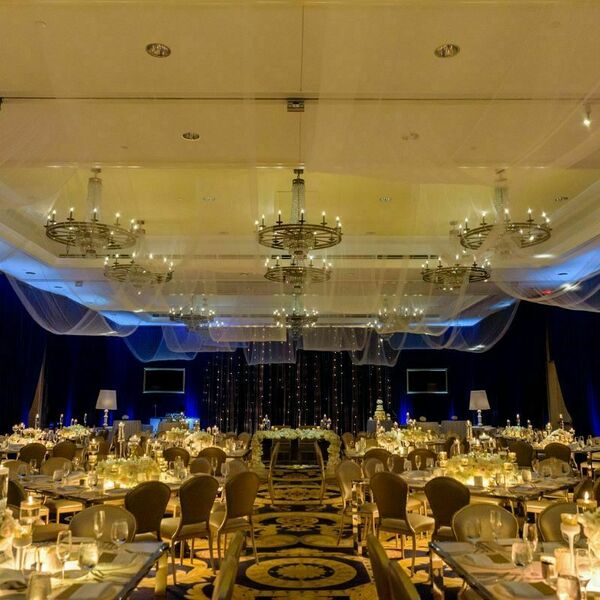 Blue velvet curtains line the sides of the room creating an intimate feel. White ribbon hangs from crystal chandeliers and string lights hang over the bride and groom table. Rectangular tables with white flowers and candles fill the room.