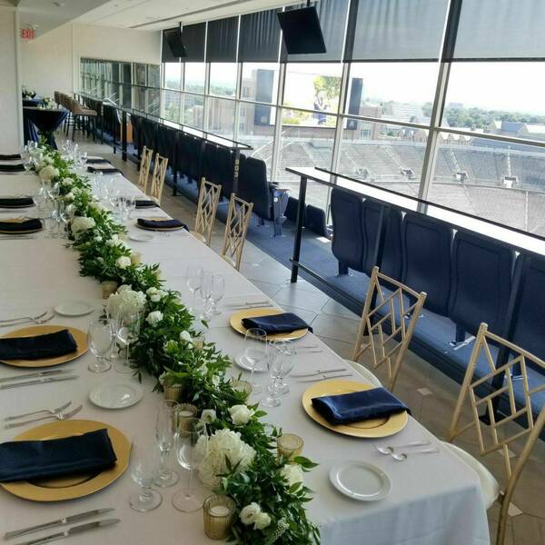 A long rectangular table decorated with a white tablecloth and gold and navy place settings fills the room. A view of the stadium can be seen in the back.