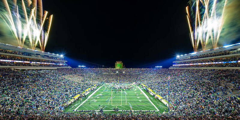 Notre Dame Stadium at night full of fans watching a football game. Celebration ensues as Notre Dame wins. Fireworks are seen on both sides of the stadium, the Hesburgh Library is lit up in green, and fans cheer.
